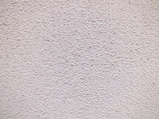 A close-up view of a textured wall with a rough, granular surface. The wall appears to be made of a material similar to stucco or concrete and has a uniform light beige color.