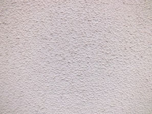 A close-up view of a textured wall with a rough, granular surface. The wall appears to be made of a material similar to stucco or concrete and has a uniform light beige color.