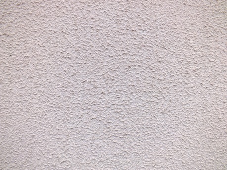 A close-up view of a textured wall with a rough, granular surface. The wall appears to be made of a material similar to stucco or concrete and has a uniform light beige color.