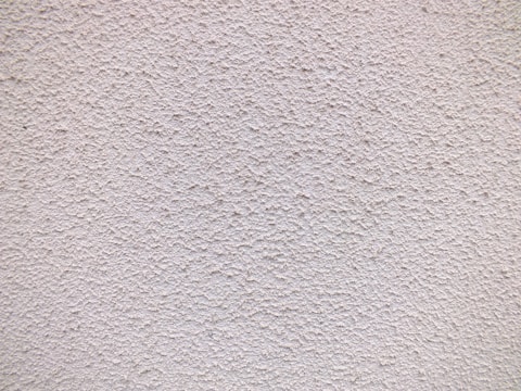 A close-up view of a textured wall with a rough, granular surface. The wall appears to be made of a material similar to stucco or concrete and has a uniform light beige color.