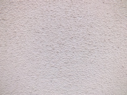 A close-up view of a textured wall with a rough, granular surface. The wall appears to be made of a material similar to stucco or concrete and has a uniform light beige color.