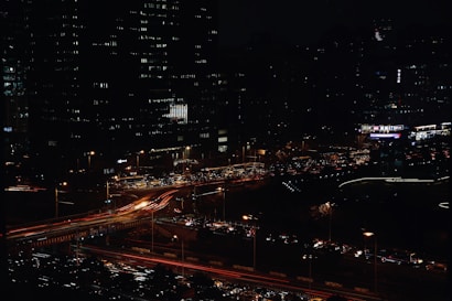 A bustling cityscape at night features bright, colorful light trails from moving vehicles on highways and streets. Tall buildings with illuminated windows tower over the traffic, creating a vibrant urban atmosphere. The dense arrangement of buildings and lights suggests a busy, modern metropolis.