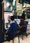Two artists are working in a small studio filled with various framed artworks and paintings. One artist is creating a detailed sketch of a man, while another artist appears to work on a different piece. The walls are adorned with a variety of paintings and decorative frames, contributing to the creative atmosphere.