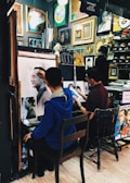 Two artists are working in a small studio filled with various framed artworks and paintings. One artist is creating a detailed sketch of a man, while another artist appears to work on a different piece. The walls are adorned with a variety of paintings and decorative frames, contributing to the creative atmosphere.