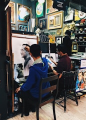 Two artists are working in a small studio filled with various framed artworks and paintings. One artist is creating a detailed sketch of a man, while another artist appears to work on a different piece. The walls are adorned with a variety of paintings and decorative frames, contributing to the creative atmosphere.