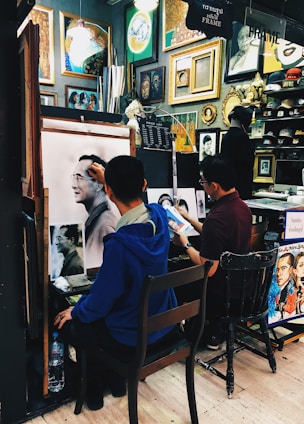 Two artists are working in a small studio filled with various framed artworks and paintings. One artist is creating a detailed sketch of a man, while another artist appears to work on a different piece. The walls are adorned with a variety of paintings and decorative frames, contributing to the creative atmosphere.