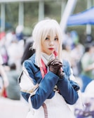 A person dressed in a detailed cosplay outfit with a blue and white costume. They have long white hair and are wearing gloves, appearing to be at an outdoor event or convention. The background is blurred, showing other people and booths.