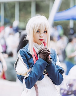 A person dressed in a detailed cosplay outfit with a blue and white costume. They have long white hair and are wearing gloves, appearing to be at an outdoor event or convention. The background is blurred, showing other people and booths.