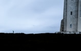 A solitary person walks near a large, stone structure like a lighthouse, with a wide expanse of cloudy sky occupying most of the frame. The mood is quiet and contemplative, enhanced by the contrast between the dark foreground and the soft, overcast sky.