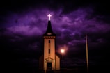 A church with a tall steeple topped by an illuminated cross stands against a dramatic, dark purple sky. A single lamp post provides soft lighting near the entrance of the church, casting subtle shadows.
