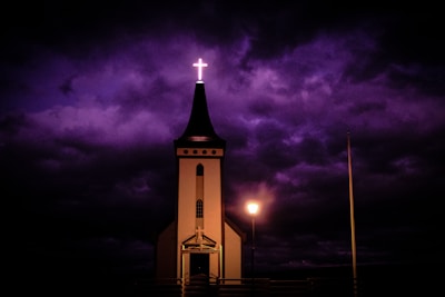 A church with a tall steeple topped by an illuminated cross stands against a dramatic, dark purple sky. A single lamp post provides soft lighting near the entrance of the church, casting subtle shadows.