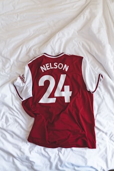 A red and white sports jersey with short sleeves is neatly laid out on a white, textured bedspread. The jersey displays the name 'Nelson' and the number '24' in bold white letters on the back.