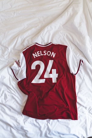 A red and white sports jersey with short sleeves is neatly laid out on a white, textured bedspread. The jersey displays the name 'Nelson' and the number '24' in bold white letters on the back.