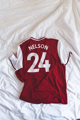 A red and white sports jersey with short sleeves is neatly laid out on a white, textured bedspread. The jersey displays the name 'Nelson' and the number '24' in bold white letters on the back.