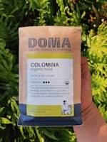 A package of Doma Coffee Roasting Company's Colombia organic huila coffee is held in someone's hand. The packaging highlights flavors of vanilla, grape, and cashew, with a medium roast level indicated. The text mentions the coffee's origin from the Aipe, Huila region of Colombia, and emphasizes its sweet flavors and syrupy body. The background features vibrant green foliage.
