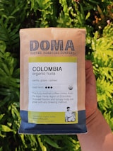 A package of Doma Coffee Roasting Company's Colombia organic huila coffee is held in someone's hand. The packaging highlights flavors of vanilla, grape, and cashew, with a medium roast level indicated. The text mentions the coffee's origin from the Aipe, Huila region of Colombia, and emphasizes its sweet flavors and syrupy body. The background features vibrant green foliage.
