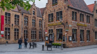 A cobblestone street scene with historic brick buildings featuring large windows and ornate architectural details. One of the buildings has a sign that reads 'Gruuthuse Hof'. There is outdoor seating with empty metal chairs and tables. Two people, one holding a camera, are captured in the center.