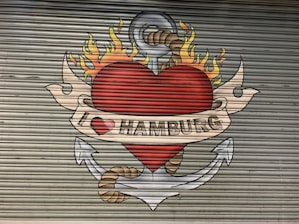 A mural painted on a corrugated metal surface depicts a large red heart with flames on top, encircled by an anchor with a rope. A beige banner with the text 'I ❤️ HAMBURG' is wrapped around the heart.
