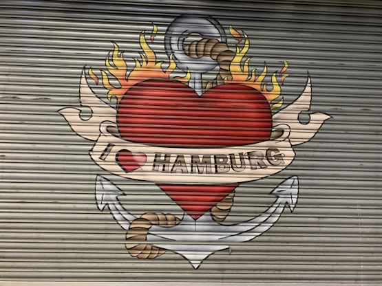 A mural painted on a corrugated metal surface depicts a large red heart with flames on top, encircled by an anchor with a rope. A beige banner with the text 'I ❤️ HAMBURG' is wrapped around the heart.