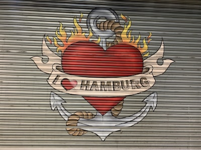 A mural painted on a corrugated metal surface depicts a large red heart with flames on top, encircled by an anchor with a rope. A beige banner with the text 'I ❤️ HAMBURG' is wrapped around the heart.