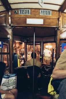 Interior view of a vintage tram showing a driver seated at the controls, navigating through a dimly lit urban street at night. The tram features wooden interior details and a classic steering wheel. Passengers are visible, one holding a newspaper, adding to the scene’s authentic feel of everyday life.