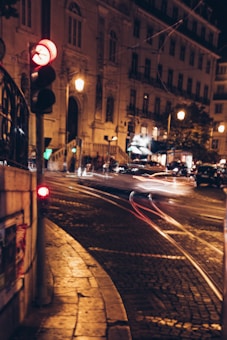 A nighttime urban scene featuring a softly-lit street corner in front of an intricately designed, historic building. Light trails from passing vehicles create a sense of motion, while pedestrians and a glowing traffic signal add life to the scene. The cobblestone street and warm lighting create a cozy, nostalgic atmosphere.