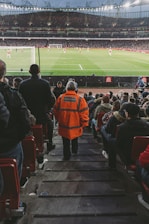 A large stadium filled with spectators, focused on a soccer match. A steward in a bright orange jacket labeled 'Arsenal Steward' stands prominently in the center of the image, overseeing the crowd. The field is visible with players actively engaged in the game.