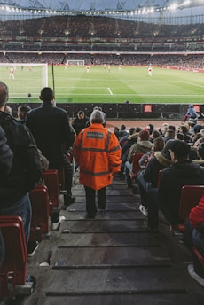 A large stadium filled with spectators, focused on a soccer match. A steward in a bright orange jacket labeled 'Arsenal Steward' stands prominently in the center of the image, overseeing the crowd. The field is visible with players actively engaged in the game.