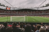A large, bustling soccer stadium filled with spectators, featuring a vivid green field under overcast skies. The focus is on a match in progress, with players visible on the pitch and a goalpost in the foreground. The stands are packed, indicating a lively atmosphere.