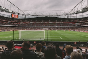 A large, bustling soccer stadium filled with spectators, featuring a vivid green field under overcast skies. The focus is on a match in progress, with players visible on the pitch and a goalpost in the foreground. The stands are packed, indicating a lively atmosphere.