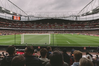 A large, bustling soccer stadium filled with spectators, featuring a vivid green field under overcast skies. The focus is on a match in progress, with players visible on the pitch and a goalpost in the foreground. The stands are packed, indicating a lively atmosphere.