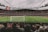 A large, bustling soccer stadium filled with spectators, featuring a vivid green field under overcast skies. The focus is on a match in progress, with players visible on the pitch and a goalpost in the foreground. The stands are packed, indicating a lively atmosphere.