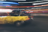 A dynamic scene features a black and yellow taxi with motion blur, suggesting speed. Neon lights streak across the background, enhancing the sense of movement and urban energy.