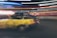 A dynamic scene features a black and yellow taxi with motion blur, suggesting speed. Neon lights streak across the background, enhancing the sense of movement and urban energy.