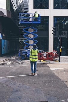 A construction worker in a bright yellow vest stands in a city street, observing two colleagues on an elevated blue scissor lift. They work on the facade of a modern building. Traffic barriers and streetlights are visible nearby, along with other urban infrastructure elements.