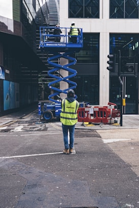 A construction worker in a bright yellow vest stands in a city street, observing two colleagues on an elevated blue scissor lift. They work on the facade of a modern building. Traffic barriers and streetlights are visible nearby, along with other urban infrastructure elements.