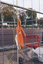 An orange safety jacket is hung on a metal fence, with construction boots on the ground nearby. There is a yellow electrical cord hanging and an orange plastic barrier visible in the background. Trees and people walking are visible in the distance beyond the barrier.
