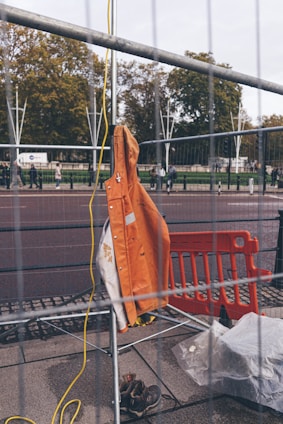 An orange safety jacket is hung on a metal fence, with construction boots on the ground nearby. There is a yellow electrical cord hanging and an orange plastic barrier visible in the background. Trees and people walking are visible in the distance beyond the barrier.