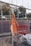 An orange safety jacket is hung on a metal fence, with construction boots on the ground nearby. There is a yellow electrical cord hanging and an orange plastic barrier visible in the background. Trees and people walking are visible in the distance beyond the barrier.