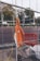 An orange safety jacket is hung on a metal fence, with construction boots on the ground nearby. There is a yellow electrical cord hanging and an orange plastic barrier visible in the background. Trees and people walking are visible in the distance beyond the barrier.