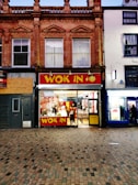 A two-story building with ornate architectural details hosts a brightly lit fast-food restaurant on the ground floor. The restaurant has large glass windows, allowing a clear view inside. People are visible, with some sitting at tables and others ordering food. The restaurant sign reads 'WOK IN' in bold yellow letters against a red background, next to a 'HALAL' sign.