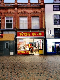 A two-story building with ornate architectural details hosts a brightly lit fast-food restaurant on the ground floor. The restaurant has large glass windows, allowing a clear view inside. People are visible, with some sitting at tables and others ordering food. The restaurant sign reads 'WOK IN' in bold yellow letters against a red background, next to a 'HALAL' sign.