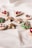 A selection of decorated Christmas cookies is arranged on a light-colored surface. The cookies come in various shapes, including stars, hearts, circles, and Christmas trees, and are adorned with icing in festive colors such as red, green, and white. Some have additional decorations like sprinkles. A red ornament is partially visible in the bottom right corner.