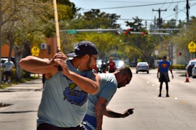 Two men appear to be engaged in a street baseball game. The man in the foreground is holding a bat and seems poised to swing. Several people are visible in the background, including one wearing a Superman t-shirt. Cars are stopped at a traffic light further down the street. The setting is urban with trees lining the sidewalks.