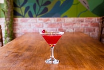 A vibrant red cocktail sits in a martini glass on a wooden table. The rim of the glass is coated with sugar, and it is garnished with a single raspberry. The background features a painted mural with leafy patterns in green and blue tones against a brick wall.