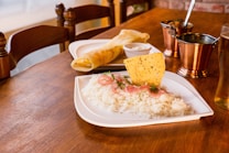 A wooden table is set with a meal featuring a plate of white rice garnished with sliced red onions, herbs, and a crispy papadum. Nearby, there is a rolled dosa with chutney. The setup includes metallic copper cups and a glass of pale beer, creating a warm, inviting dining atmosphere.