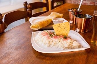 A wooden table is set with a meal featuring a plate of white rice garnished with sliced red onions, herbs, and a crispy papadum. Nearby, there is a rolled dosa with chutney. The setup includes metallic copper cups and a glass of pale beer, creating a warm, inviting dining atmosphere.