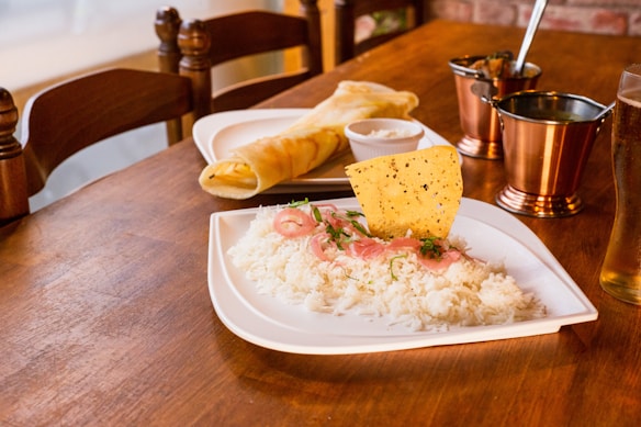 A wooden table is set with a meal featuring a plate of white rice garnished with sliced red onions, herbs, and a crispy papadum. Nearby, there is a rolled dosa with chutney. The setup includes metallic copper cups and a glass of pale beer, creating a warm, inviting dining atmosphere.