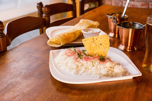A wooden table is set with a meal featuring a plate of white rice garnished with sliced red onions, herbs, and a crispy papadum. Nearby, there is a rolled dosa with chutney. The setup includes metallic copper cups and a glass of pale beer, creating a warm, inviting dining atmosphere.