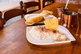 A wooden table is set with a meal featuring a plate of white rice garnished with sliced red onions, herbs, and a crispy papadum. Nearby, there is a rolled dosa with chutney. The setup includes metallic copper cups and a glass of pale beer, creating a warm, inviting dining atmosphere.